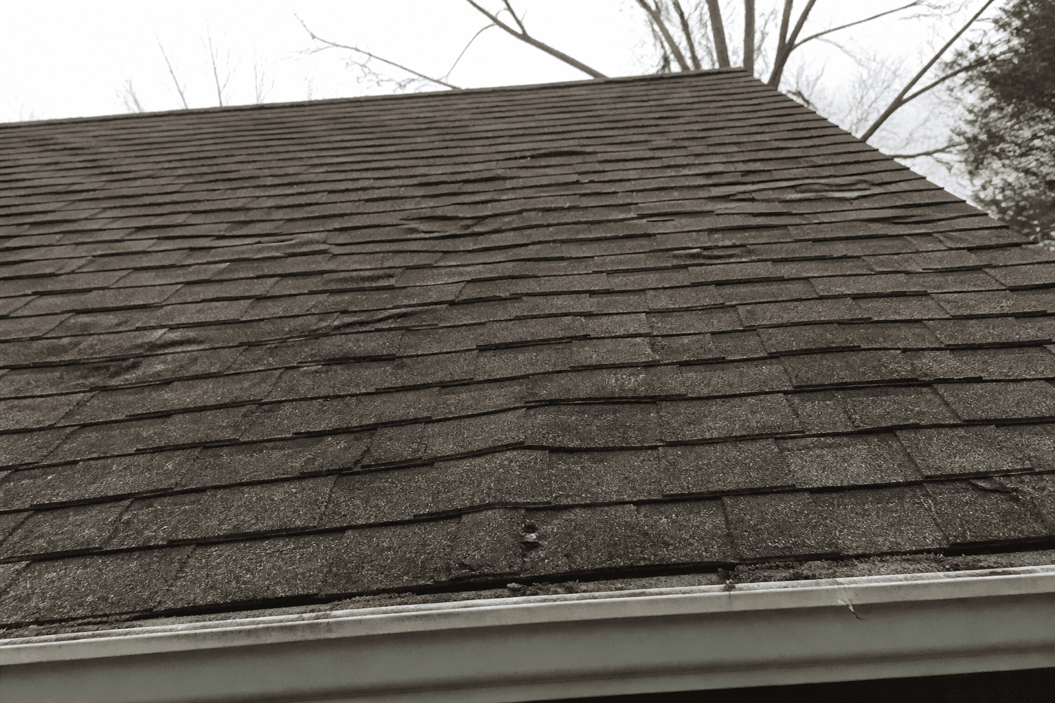 Weathered brown-gray shingle slope with wavy wrinkled courses, algae streaks, and a gutter at the eave