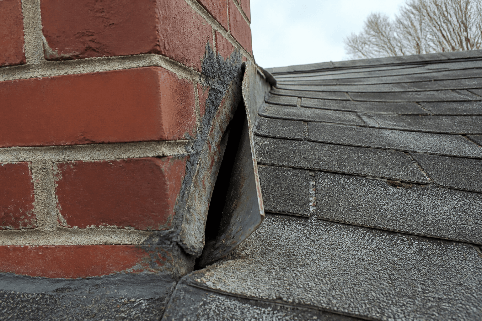 Brick chimney on an aged shingle roof with lifted rusted step flashing and cracked sealant at the joint