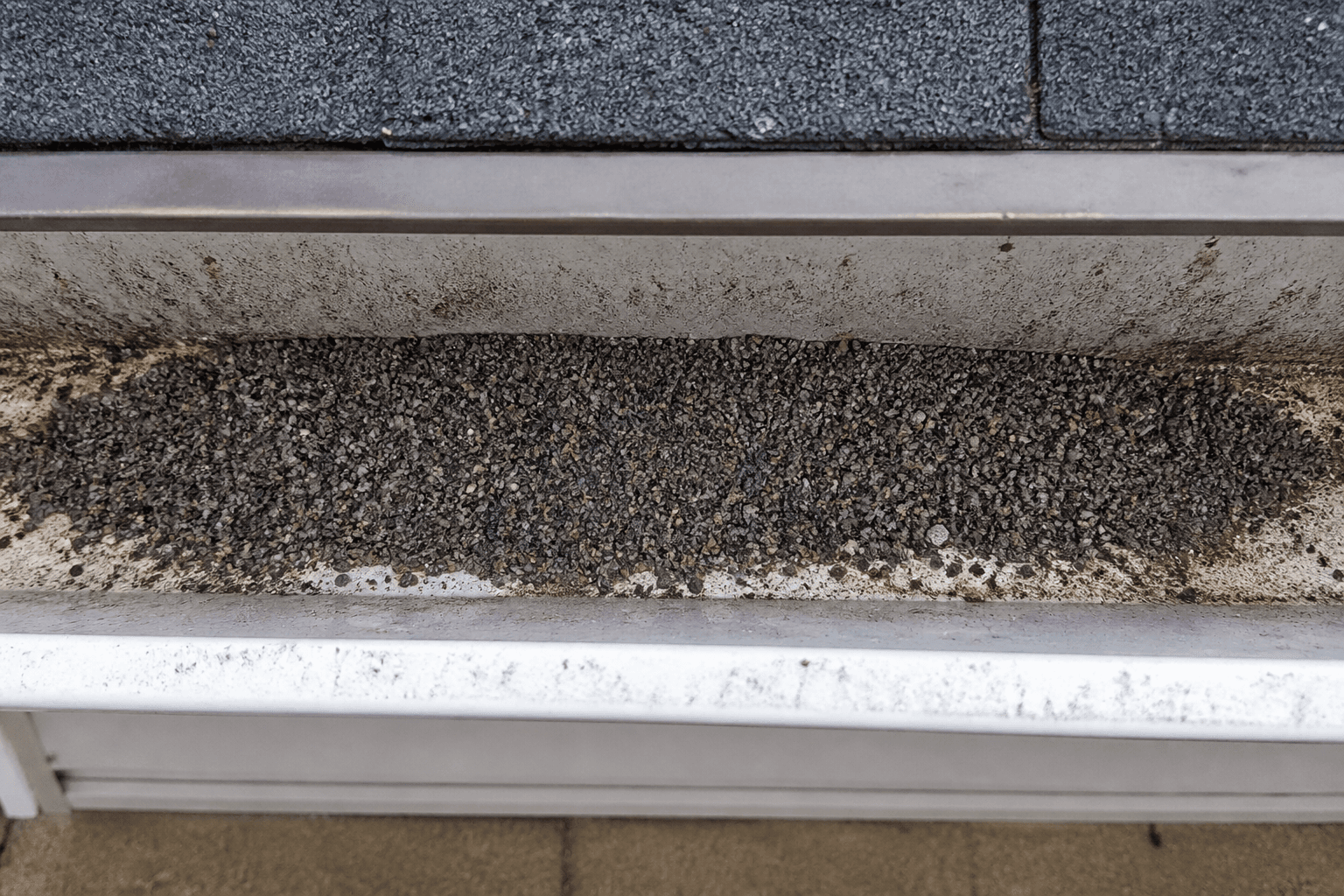 White rain gutter filled with a pile of dark asphalt shingle granules, drip edge and shingles visible above
