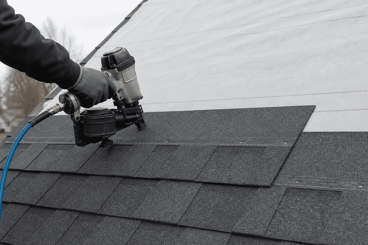 Nail gun fastening a charcoal architectural shingle; gray underlayment still open above the last course