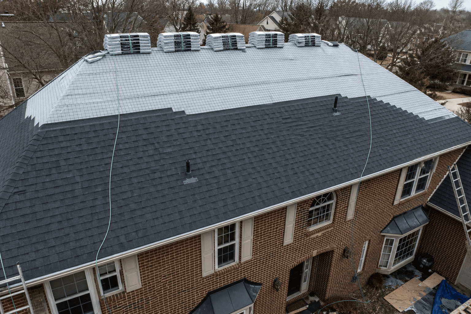 House roof in progress: new charcoal shingles on the lower slope, gray underlayment still open toward the ridge, bundles at the peak